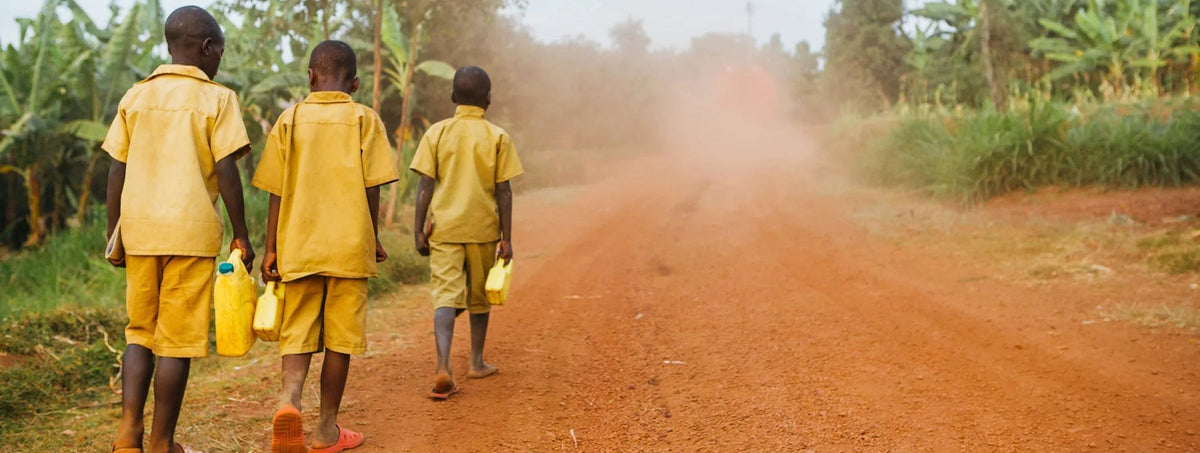 Three children in yellow uniforms walking down a dusty road carrying yellow water containers.