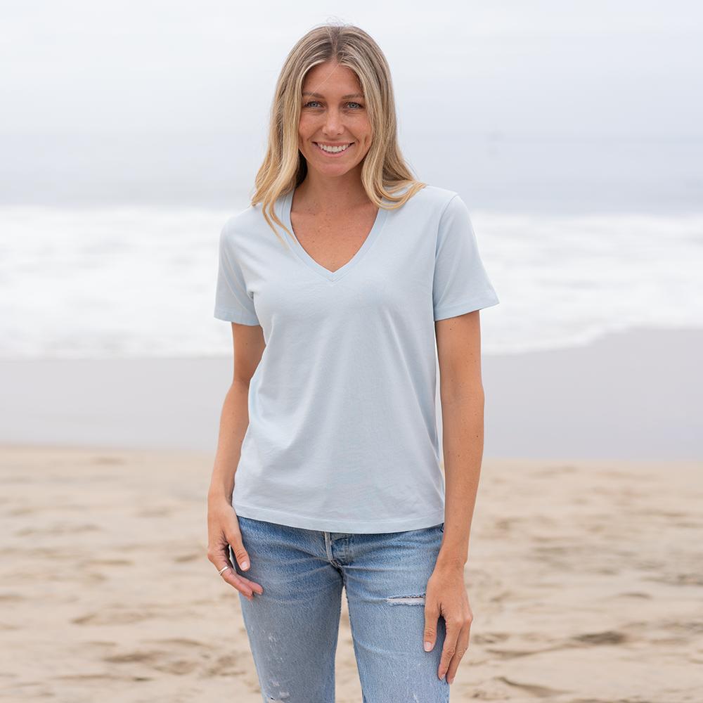 Woman wearing a light blue organic cotton V-neck t-shirt and jeans on a sandy beach.